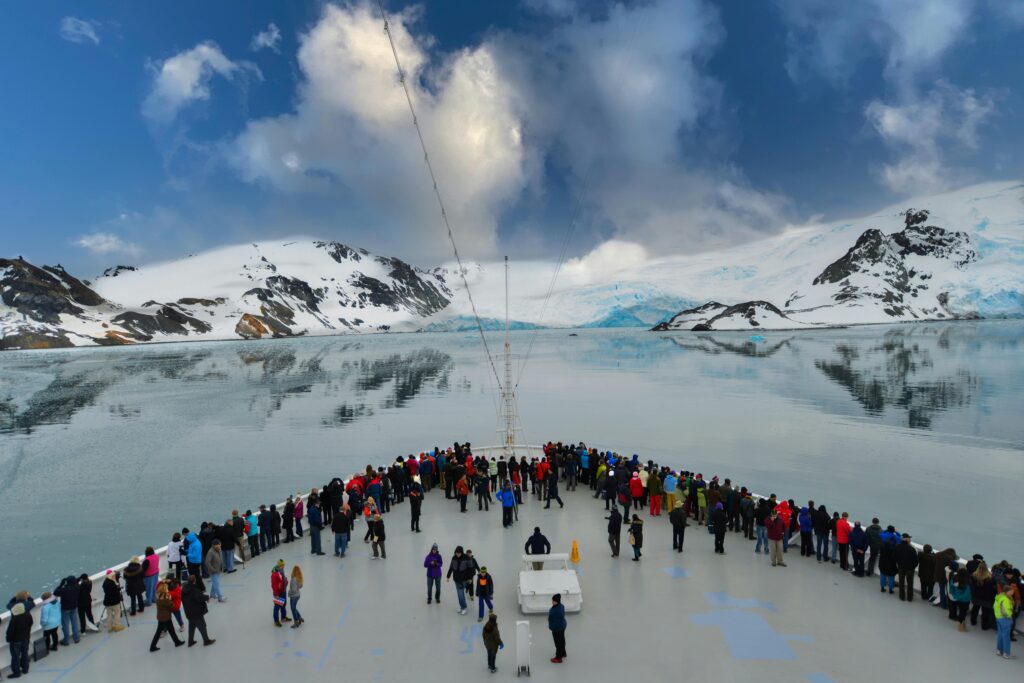 A cruise ship deck filled with people enjoying stunning views of Antarctic icebergs and snow-capped mountains.