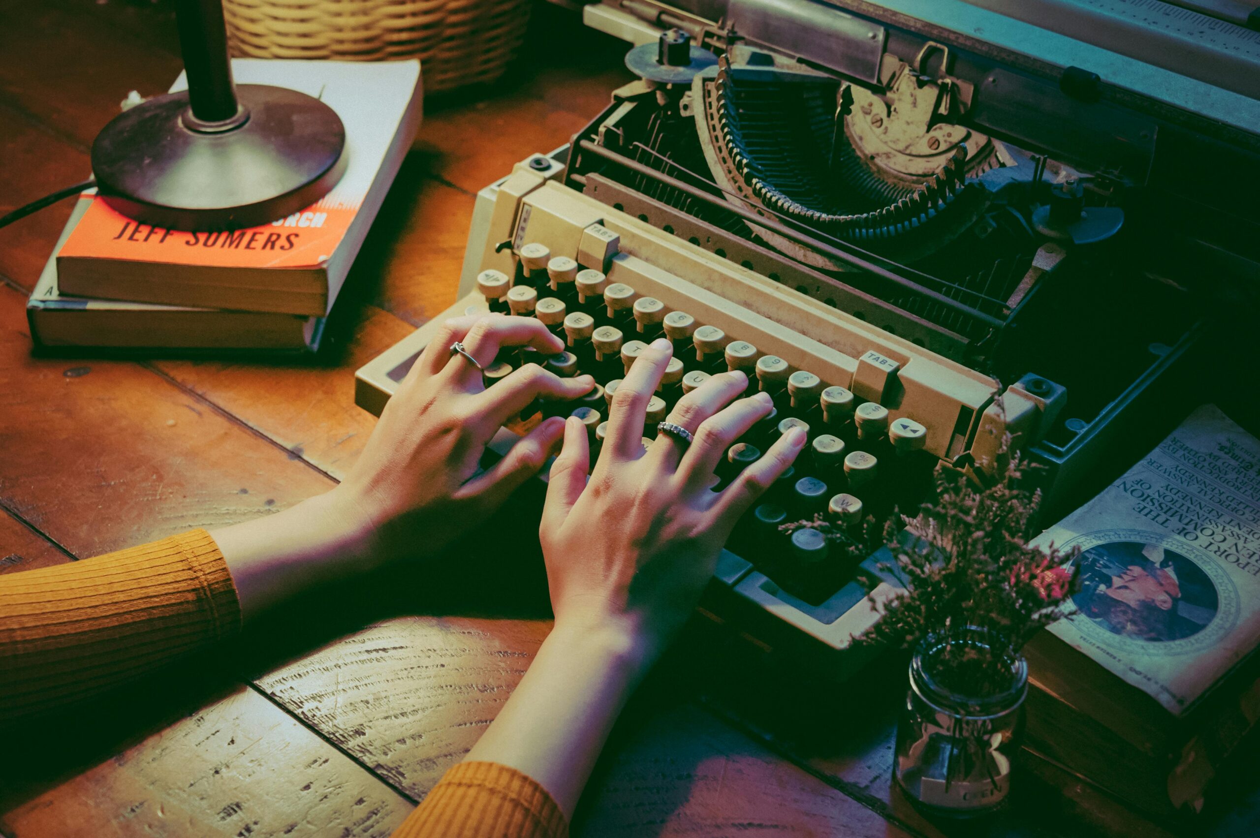 A cozy scene with a vintage typewriter and hands typing indoors beside books and flowers.