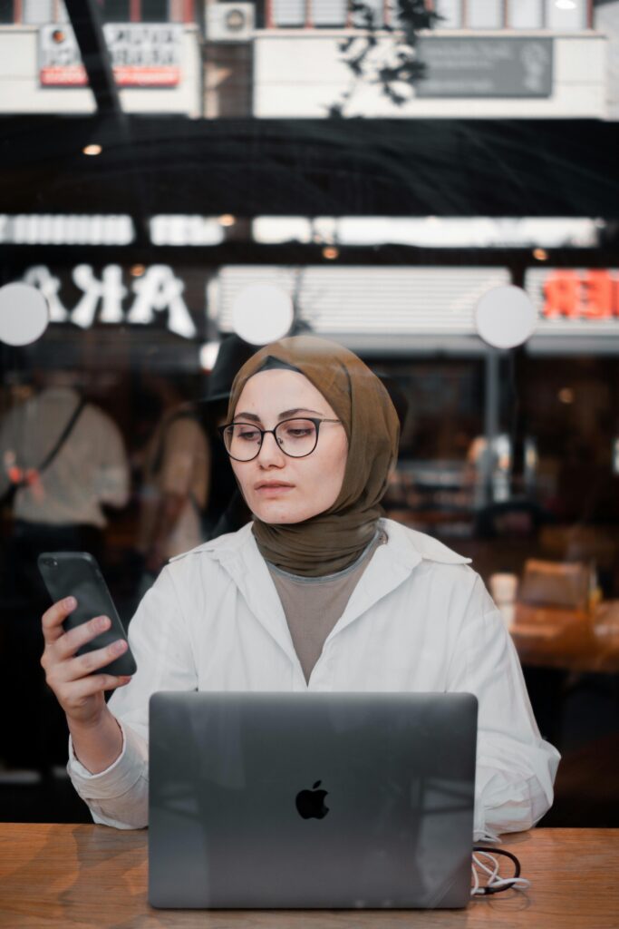 Middle Eastern woman using smartphone and laptop in café.