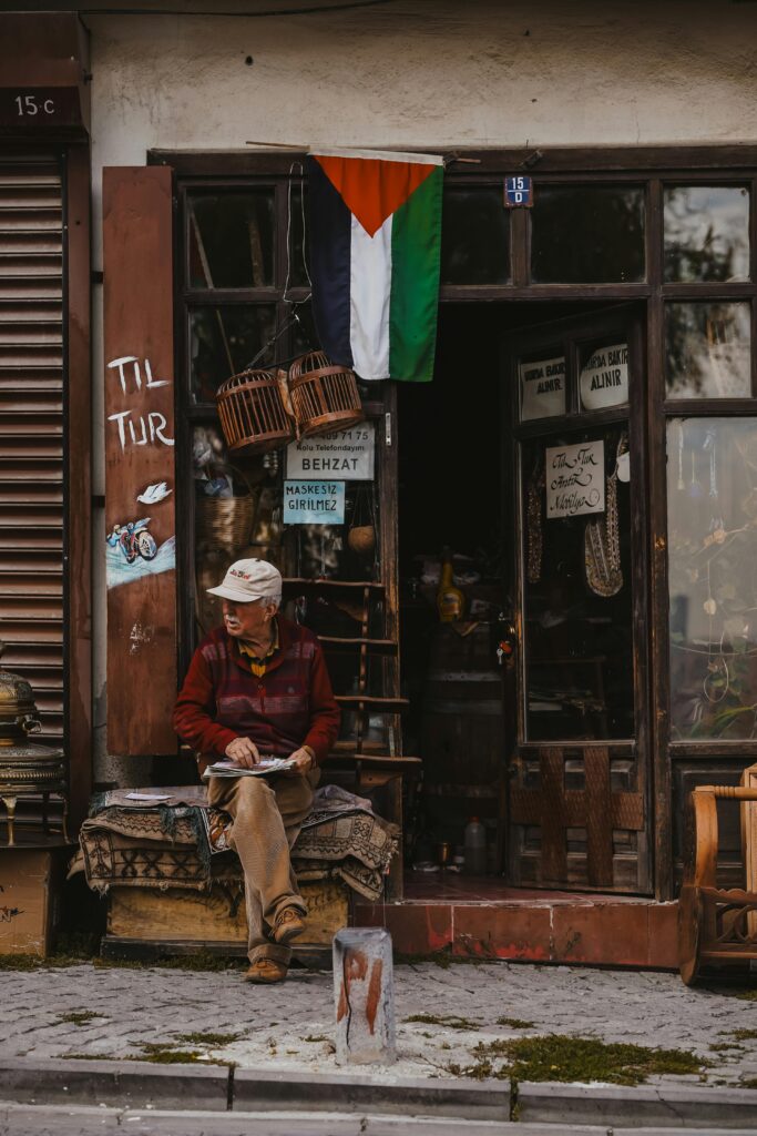 Elderly Man Sitting in Front of a Store with a Palestinian Flag Above the Entrance