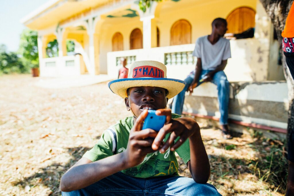 A young boy enjoys leisure time outdoors in Haiti, wearing a straw hat and holding a phone.