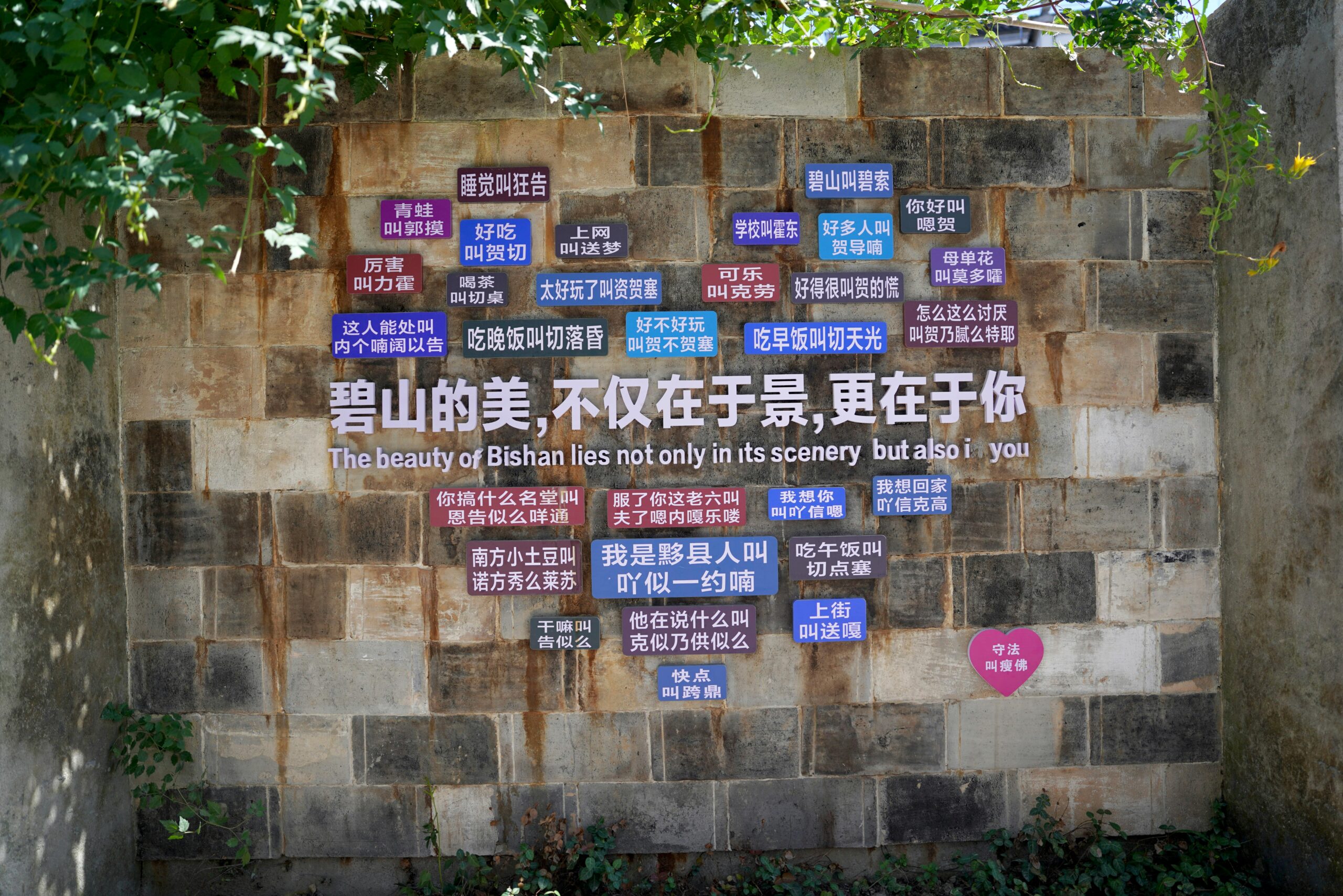 Urban wall with motivational quote in multiple languages, surrounded by greenery.