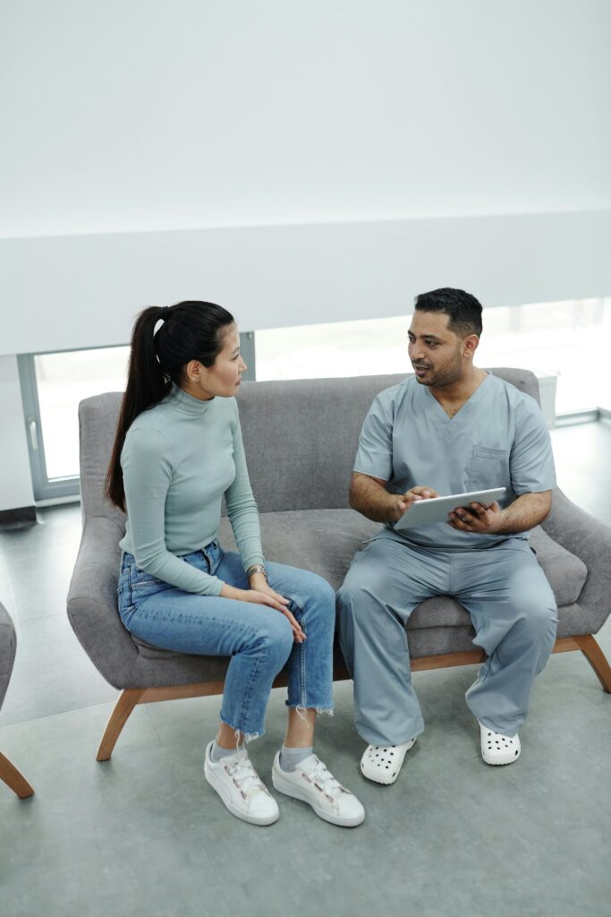 A doctor in scrub suit consults a patient on a couch using a tablet indoors.