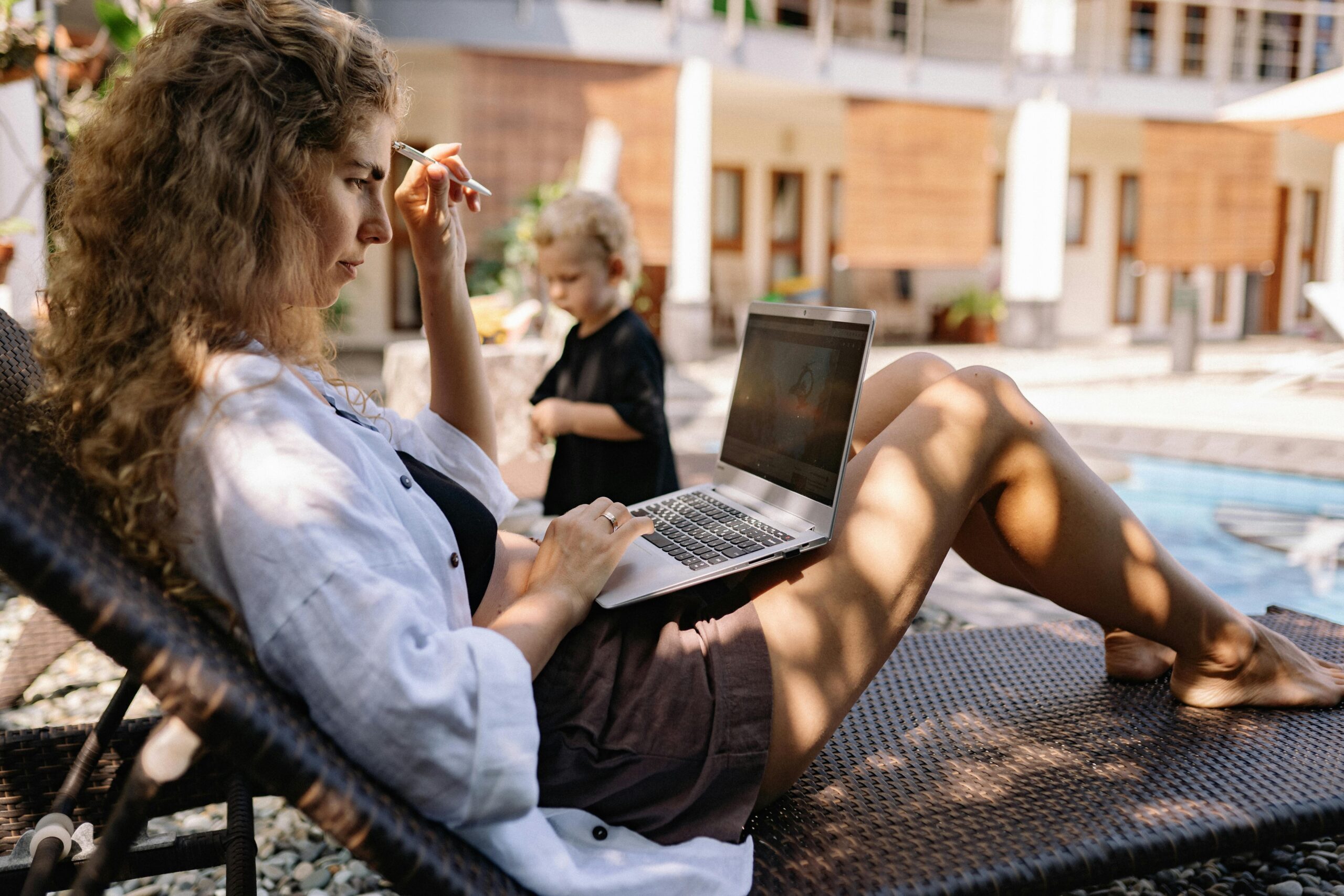 Woman typing on a laptop while relaxing outdoors on a sunlounger by the pool.