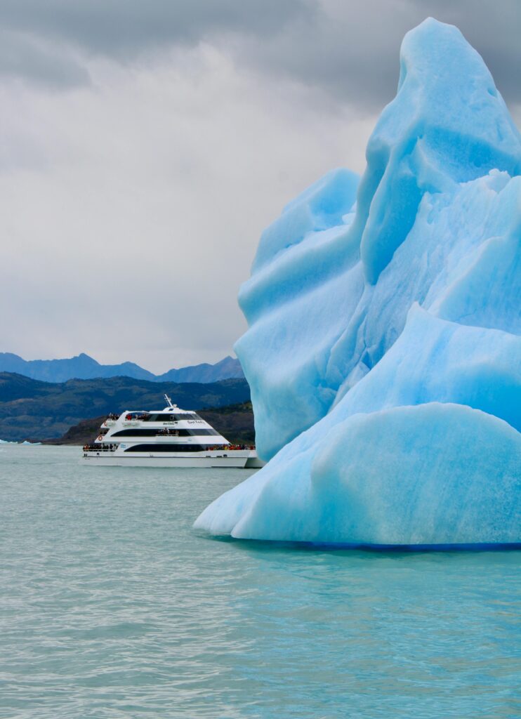 A cruise ship cruises past a large iceberg in Arctic waters, with mountains in the background.