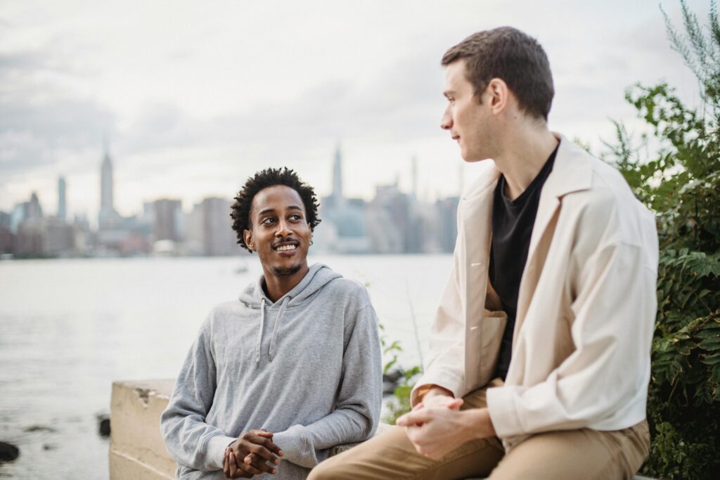 Positive African American male leaning on concrete border while having conversation with friend on city waterfront