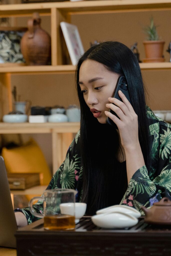 Asian woman working remotely on a laptop while talking on the phone and enjoying tea.