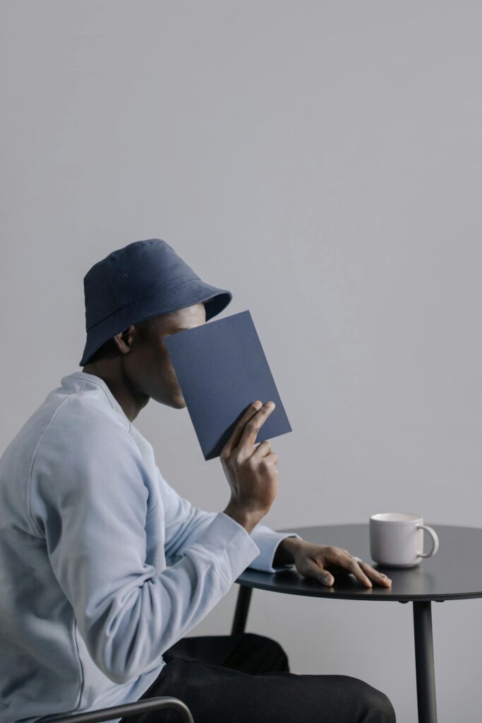 A man in a blue bucket hat reads a book at a table indoors, creating a serene and thoughtful atmosphere.