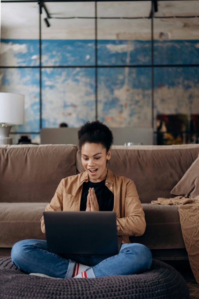 A young woman seated on the floor works enthusiastically on her laptop indoors.