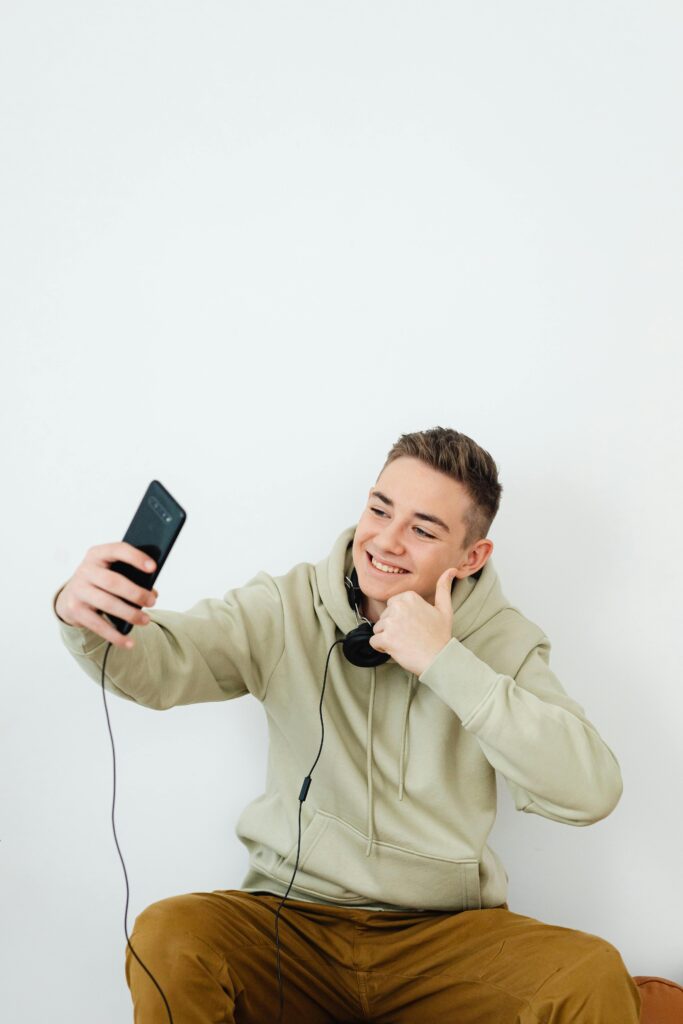 Young man in hoodie makes a thumbs-up gesture while taking a selfie, wearing headphones.