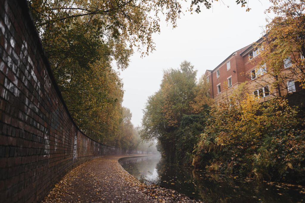 Narrow water channel flowing between sidewalk with fallen leaves near brick wall and residential house in town on foggy autumn day