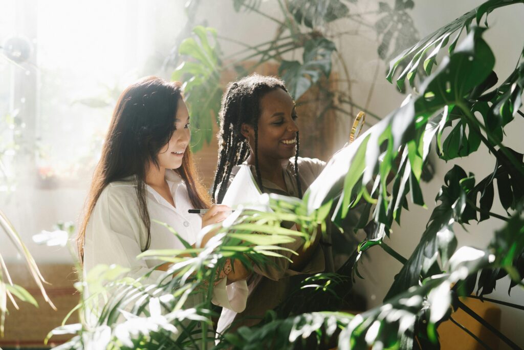 Two women botanists examining monstera plants indoors with sunlight filtering through leaves.