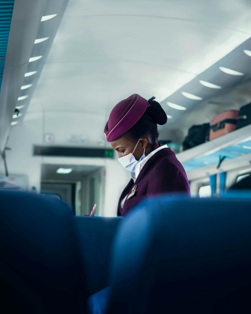 A flight attendant in a purple uniform wearing a surgical mask, attending to passengers in an airplane cabin.