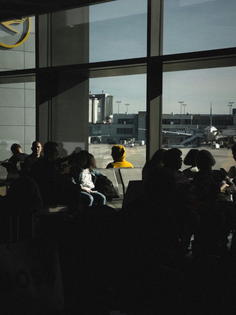 Silhouetted travelers in an airport terminal waiting area near large windows with outside tarmac view.