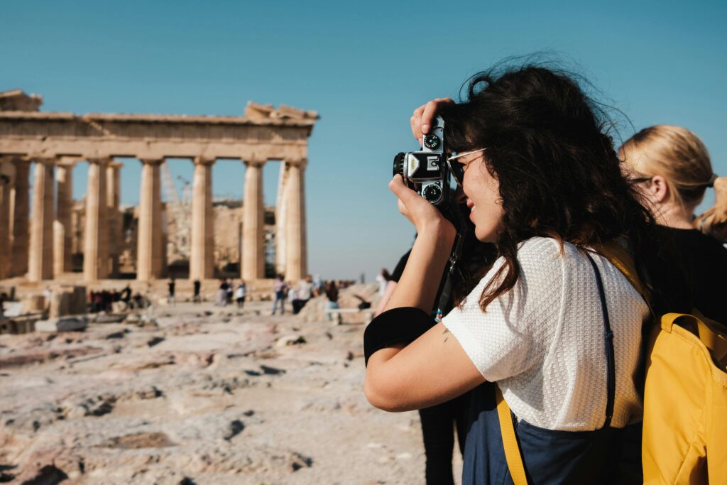 A woman captures an image of the iconic Parthenon in Athens with a vintage camera on a sunny day.