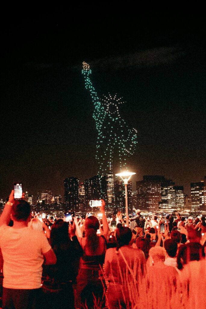 A vibrant drone light show forms the Statue of Liberty as a crowd captures the spectacle at night.