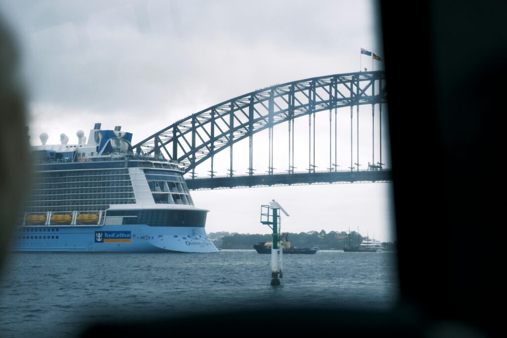 Cruise ship sailing under the iconic Sydney Harbor Bridge on a cloudy day.