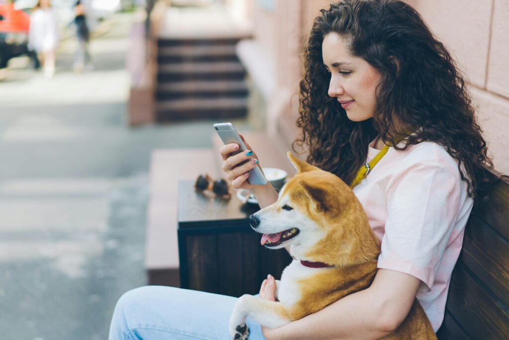 A woman sitting on a bench with a dog, using her smartphone in an urban setting.