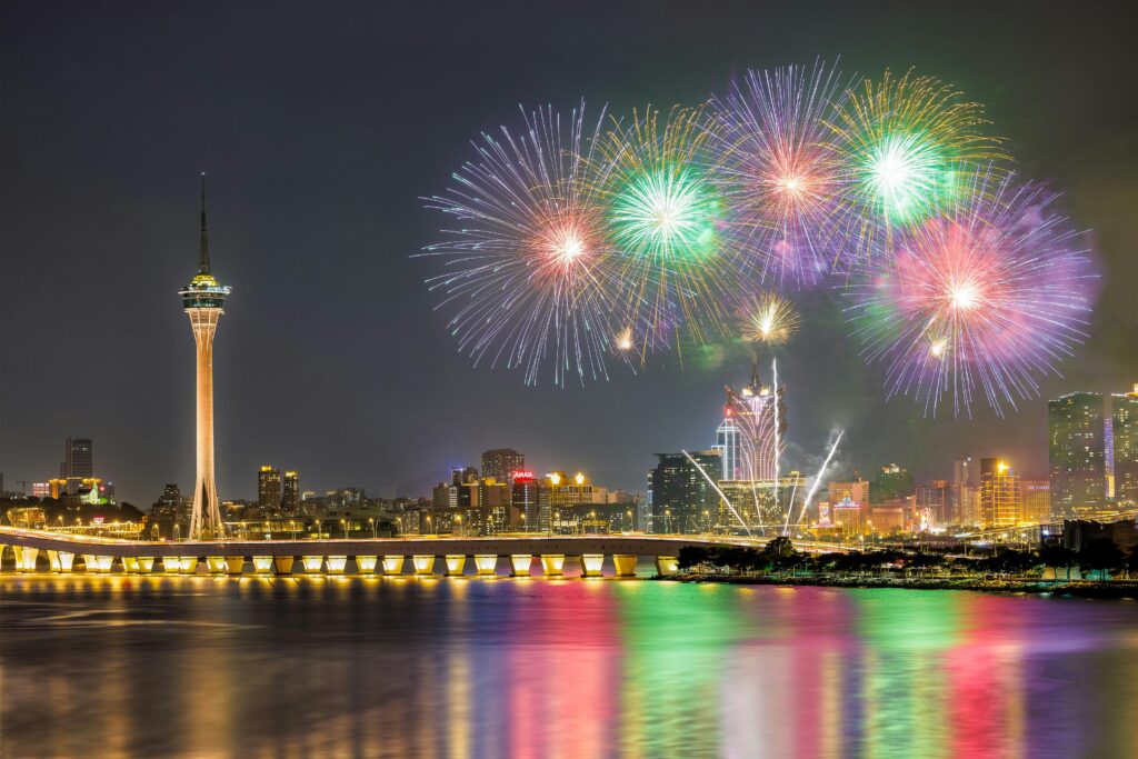 Vibrant fireworks illuminate the skyline of Macau with bright colors reflecting on the water at night.