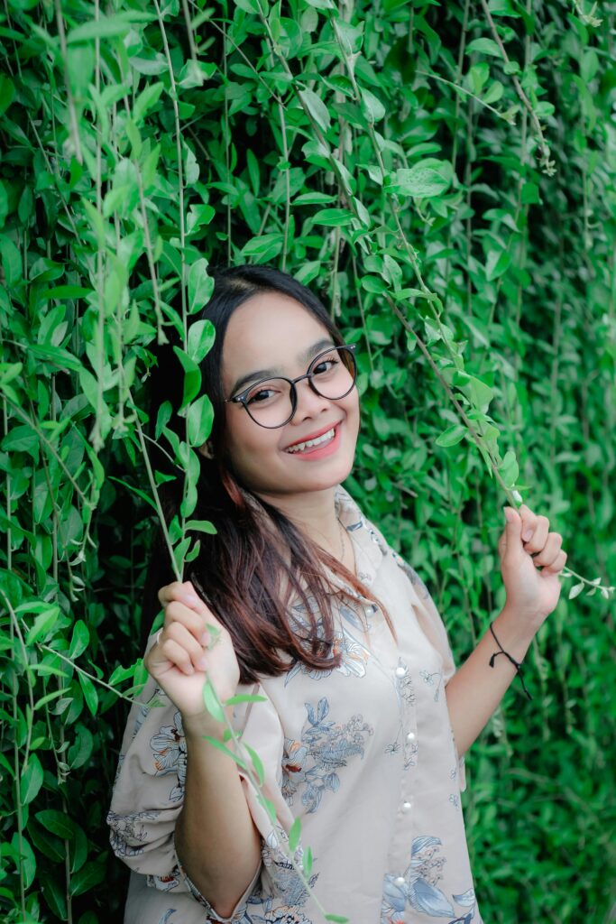 Cheerful woman with glasses standing among vibrant green foliage outdoors.