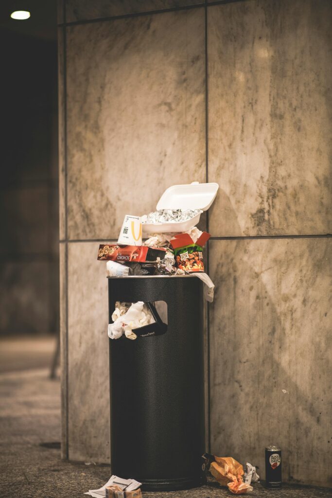 A trash can overflowing with garbage in an urban setting, against a concrete wall.