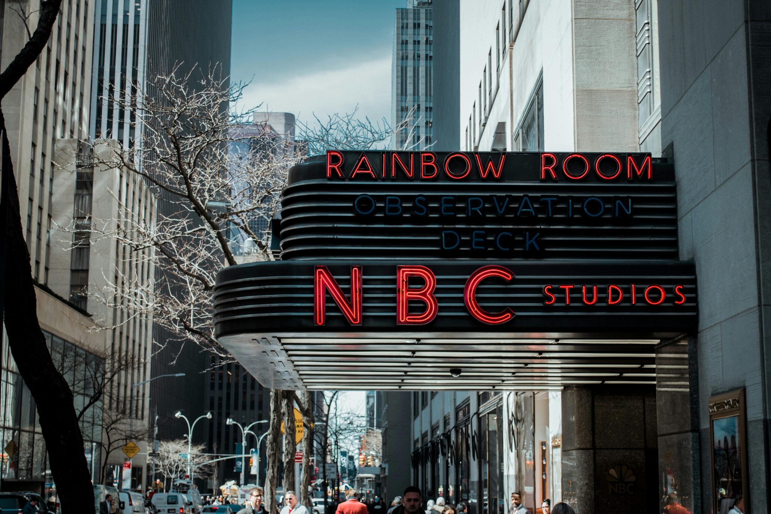 A busy urban scene in New York City featuring the iconic NBC Studios sign on a sunny day.