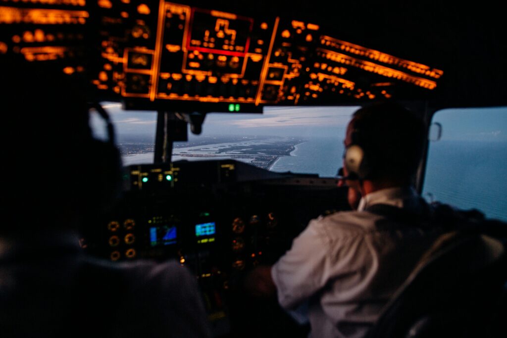 Pilots operating an aircraft cockpit during evening flight, showing illuminated control panels and distant coastline.