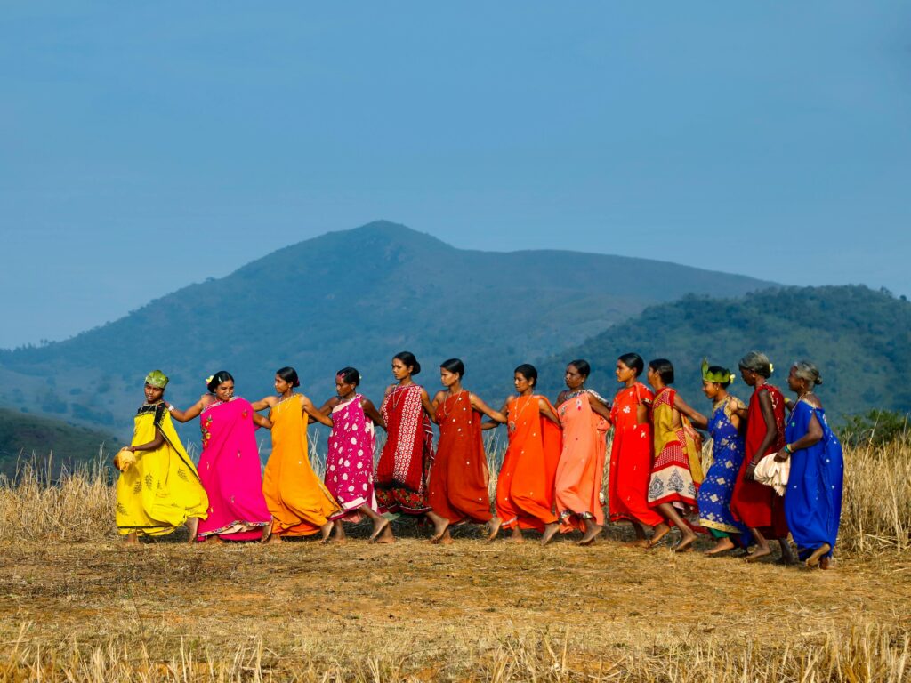 A group of women perform a traditional tribal dance in vibrant clothing against a rural Indian backdrop.