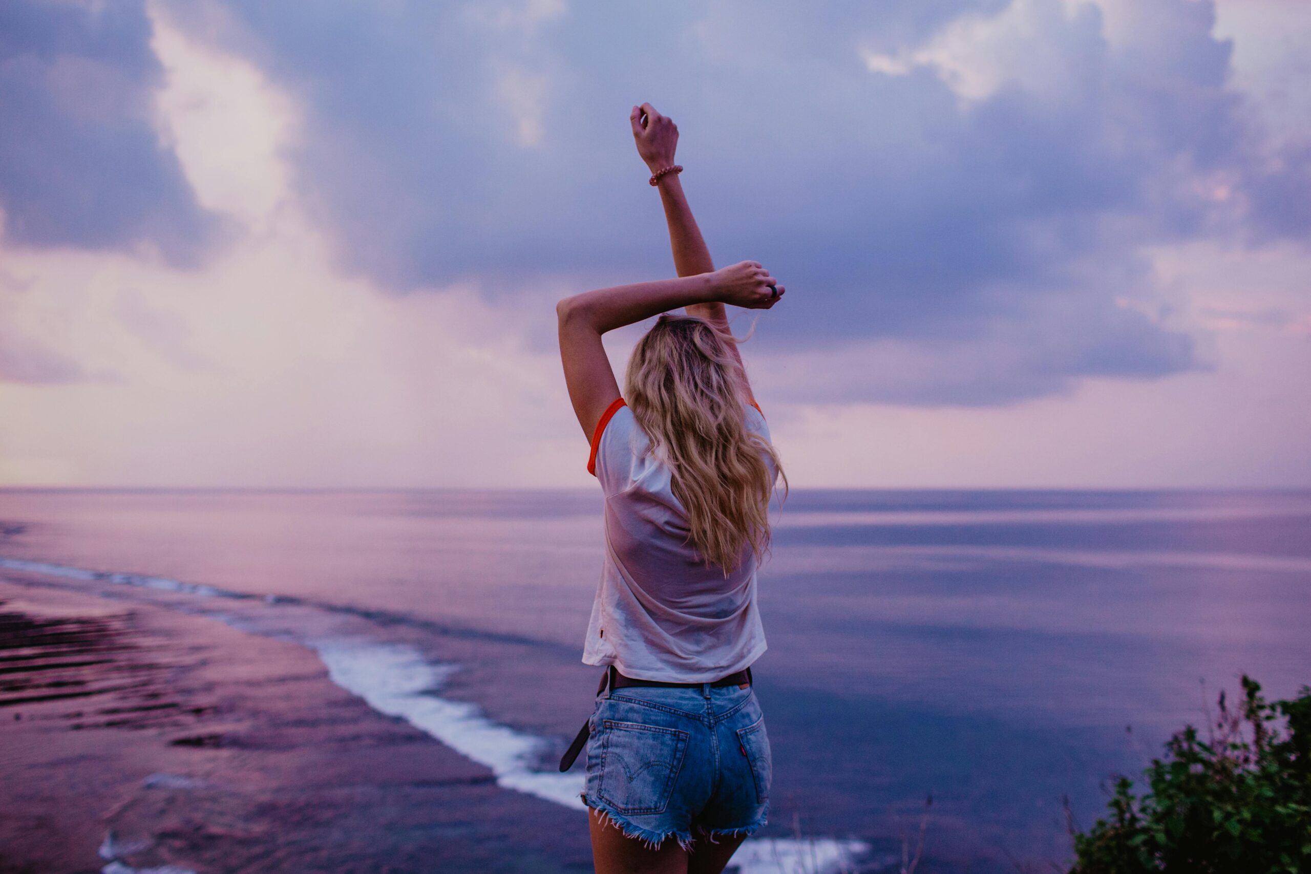 Back view anonymous female in denim shorts and white top standing on sandy beach with arms raised and enjoying picturesque view of evening seascape and purple sky