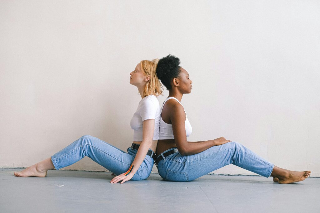 Two diverse women sit back to back in matching outfits, highlighting contrast and unity.