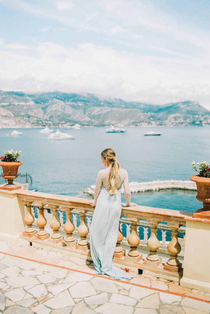 Blonde woman in a long dress on a terrace, gazing at the sea with mountains in the background.