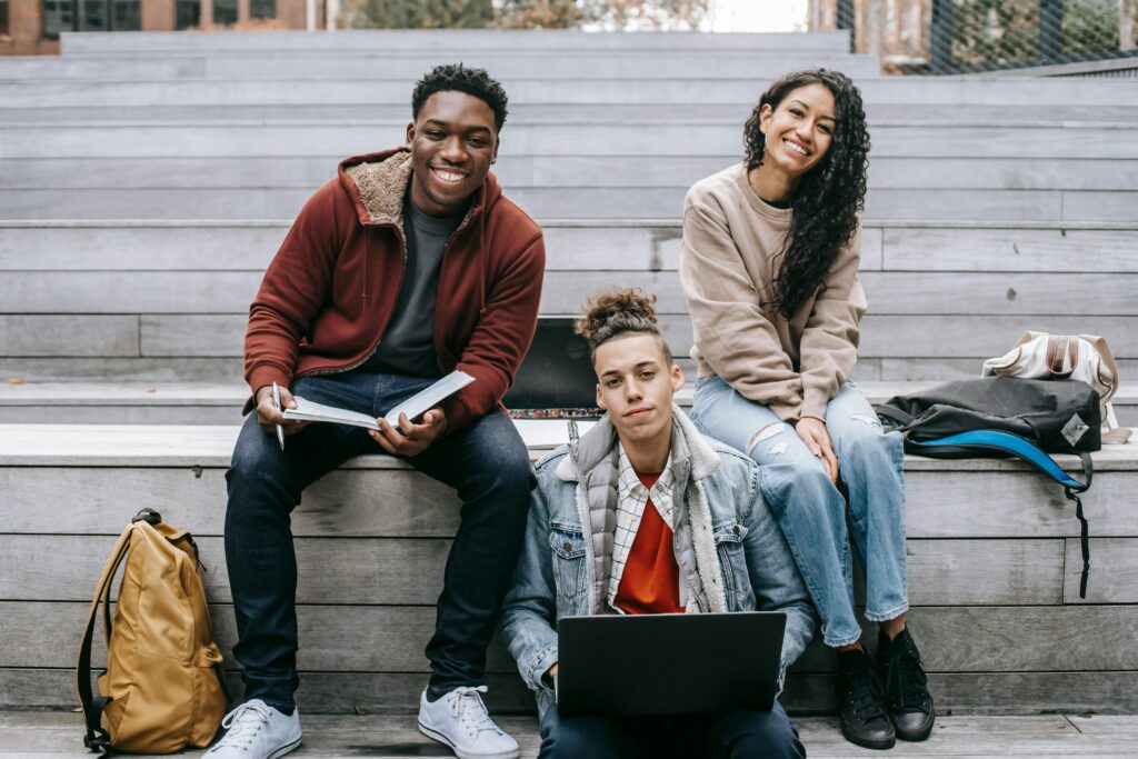 Three young adults with laptops studying together outdoors with cheerful expressions.