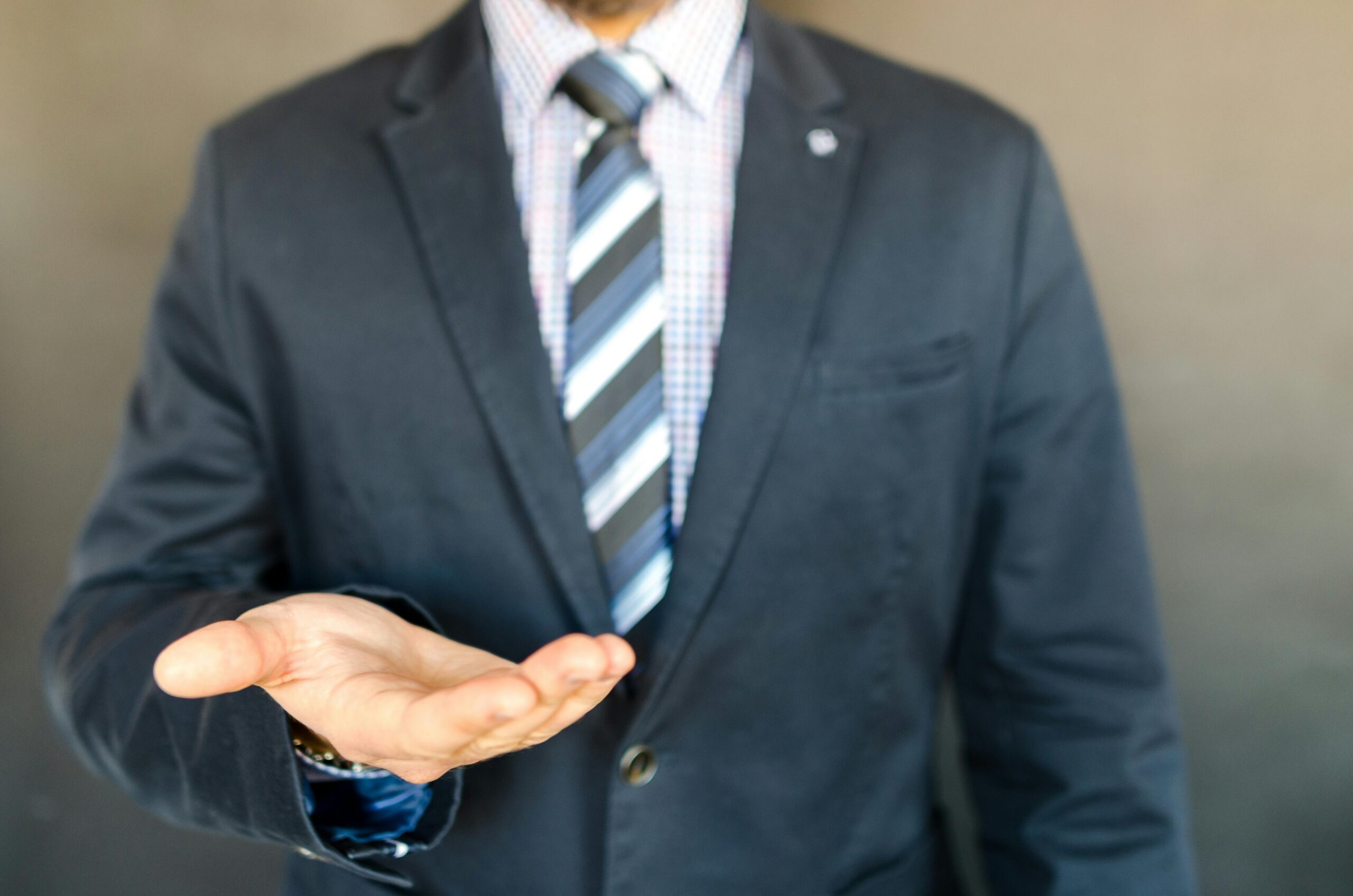 Businessman in a suit gestures with open palm, indoors, conveying offering or asking.