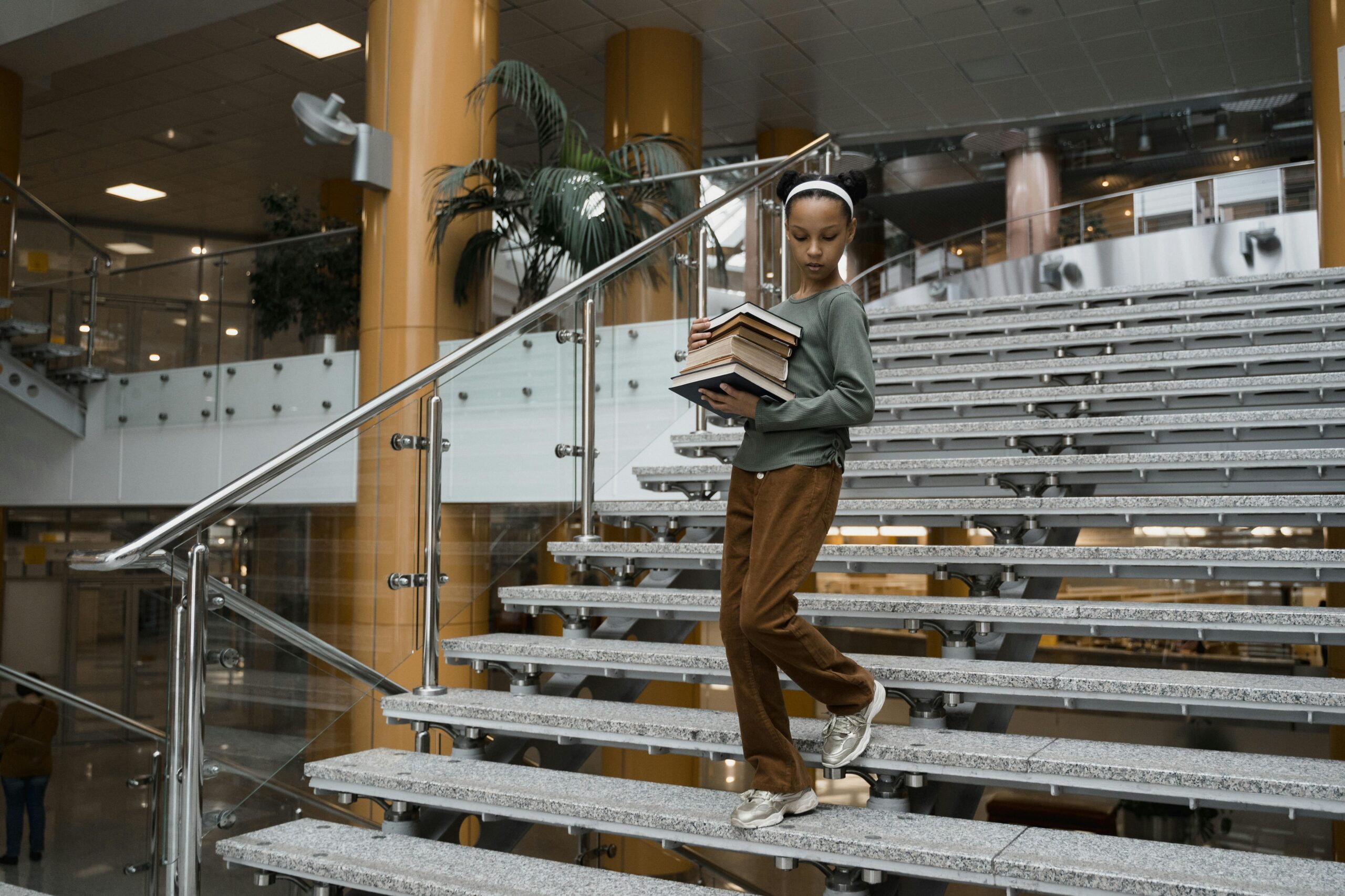 African American girl descends stairs with a stack of books in a modern library setting.