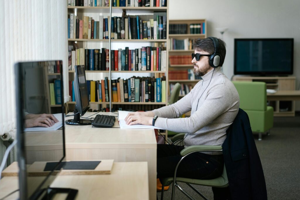 A blind man uses a computer with headphones in a library, highlighting technology's role in accessibility.