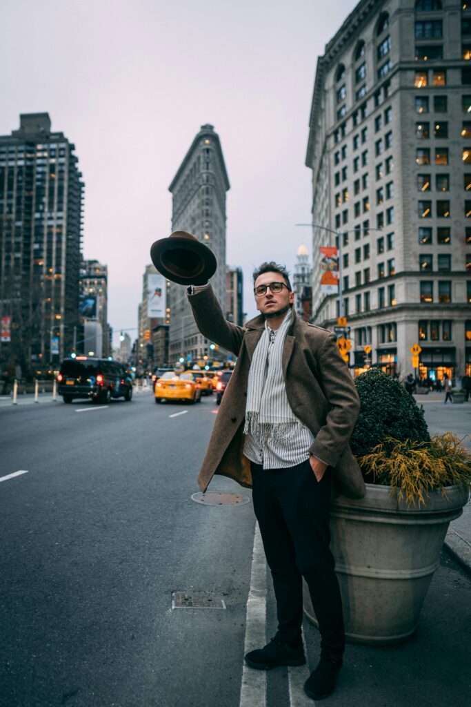 Man in coat waving hat on a busy New York City street with the Flatiron Building in view.