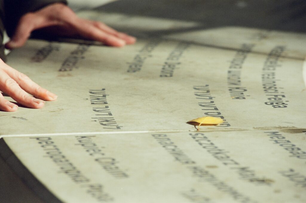 Close-up of hands on engraved stone with yellow leaf. Text in various languages. Autumn scene.