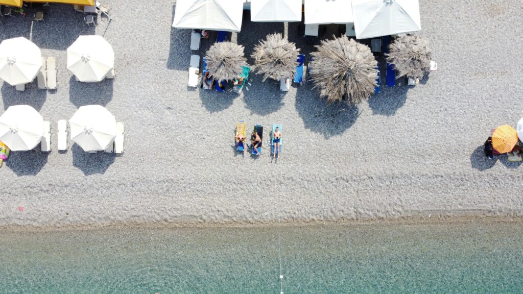 Drone shot of Kiato beach in Greece with umbrellas, lounge chairs, and sunbathers enjoying summer.