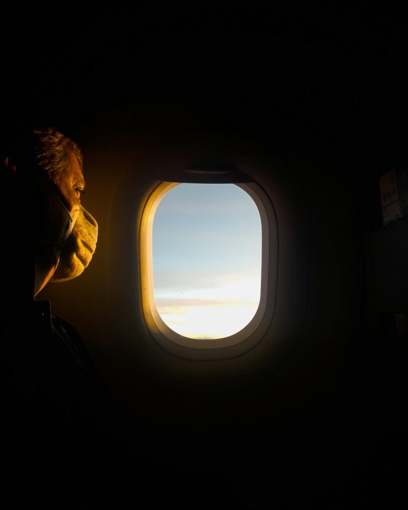 Silhouette of a masked passenger gazing at dawn sky through airplane window.