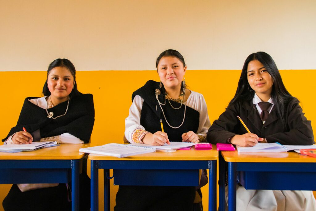 Three students in traditional attire sit at desks, engaged in classroom activities.