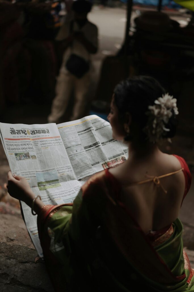 A woman in a sari reads a newspaper outdoors, captured from behind.