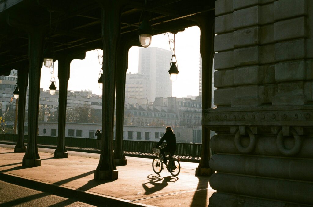 A person bikes under an iron bridge in Paris, silhouetted by the sunrise, showcasing urban architecture.