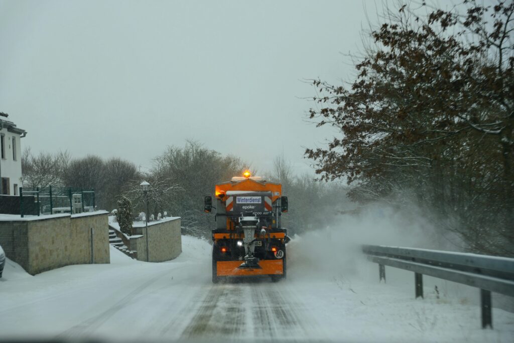 Orange snowplow clears a rural road during winter snowfall.