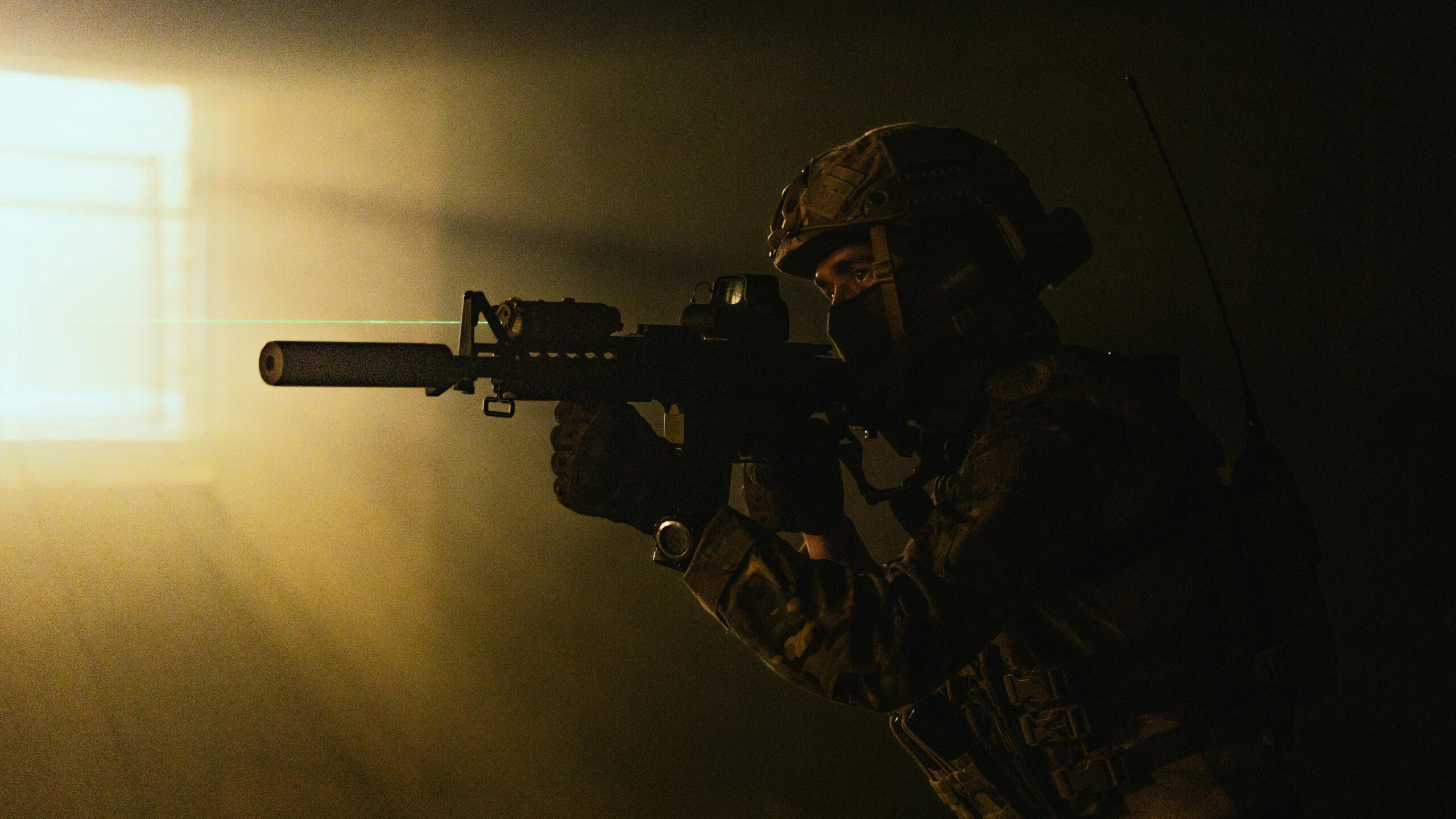 A soldier in tactical gear aims a weapon with a laser sight in low light conditions.