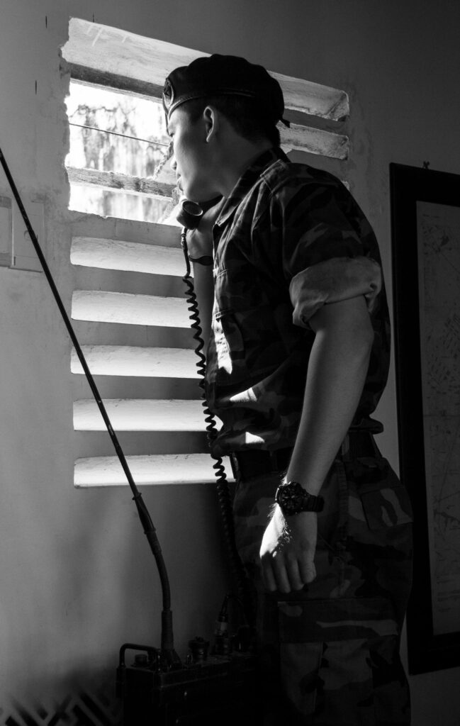 A soldier in camouflage uniform using a radio phone while looking through a window.