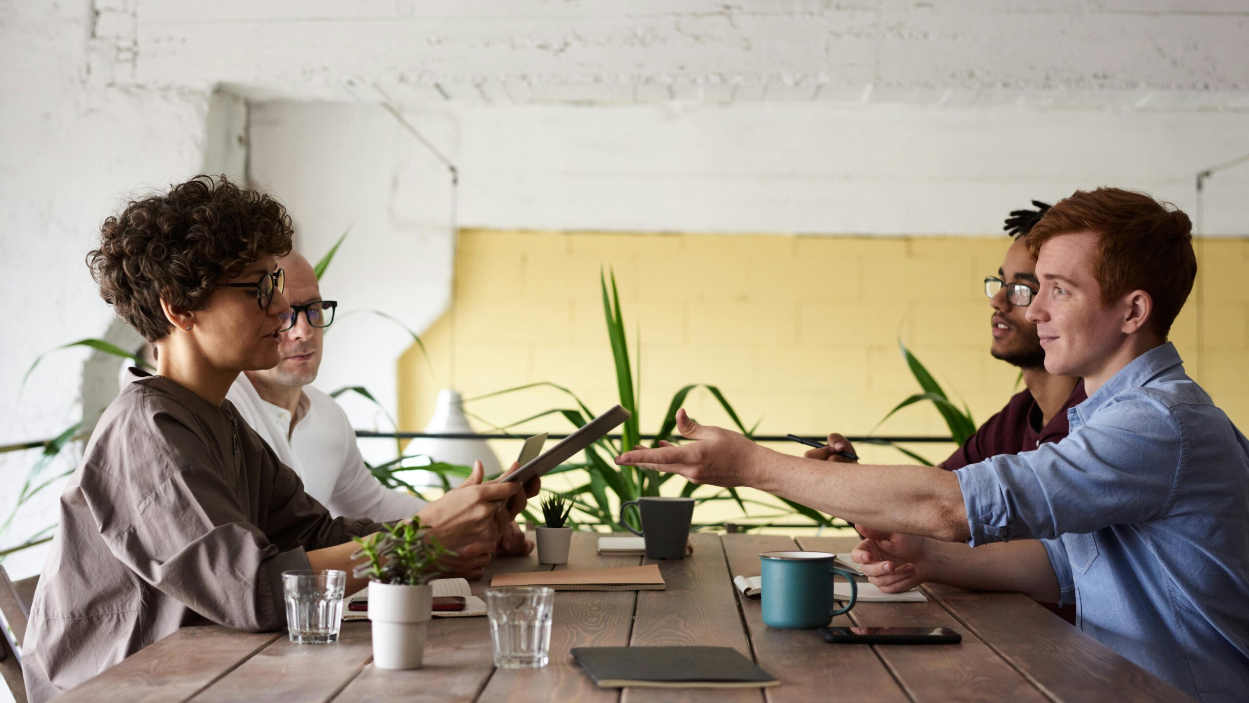 A diverse team of professionals having a collaborative meeting in an indoor setting with plants and coffee mugs.