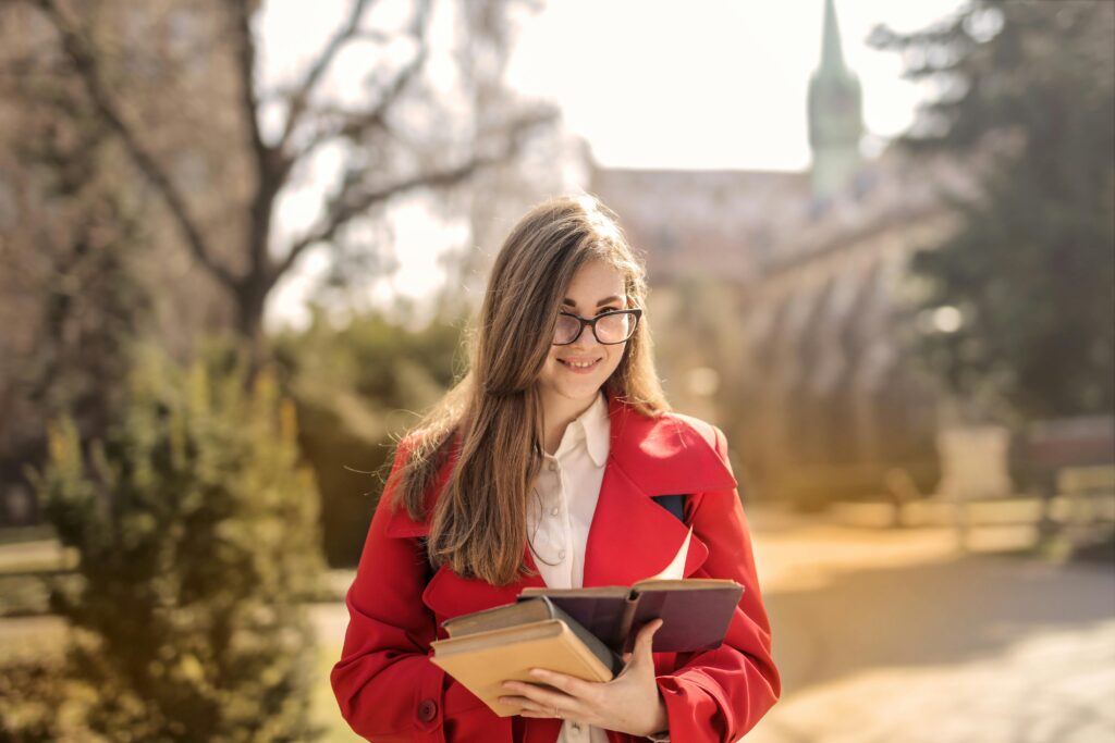 Smiling young woman in red coat holding books outdoors with a soft focus background.