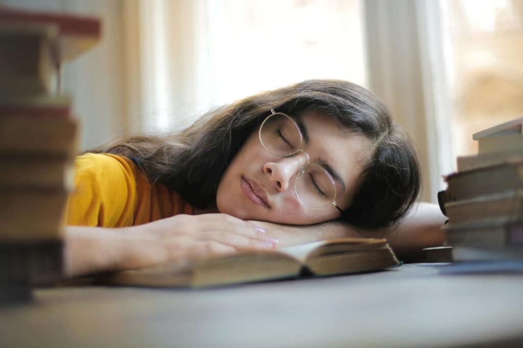 A young woman with glasses naps on a book indoors during the day, capturing a moment of relaxation and fatigue.