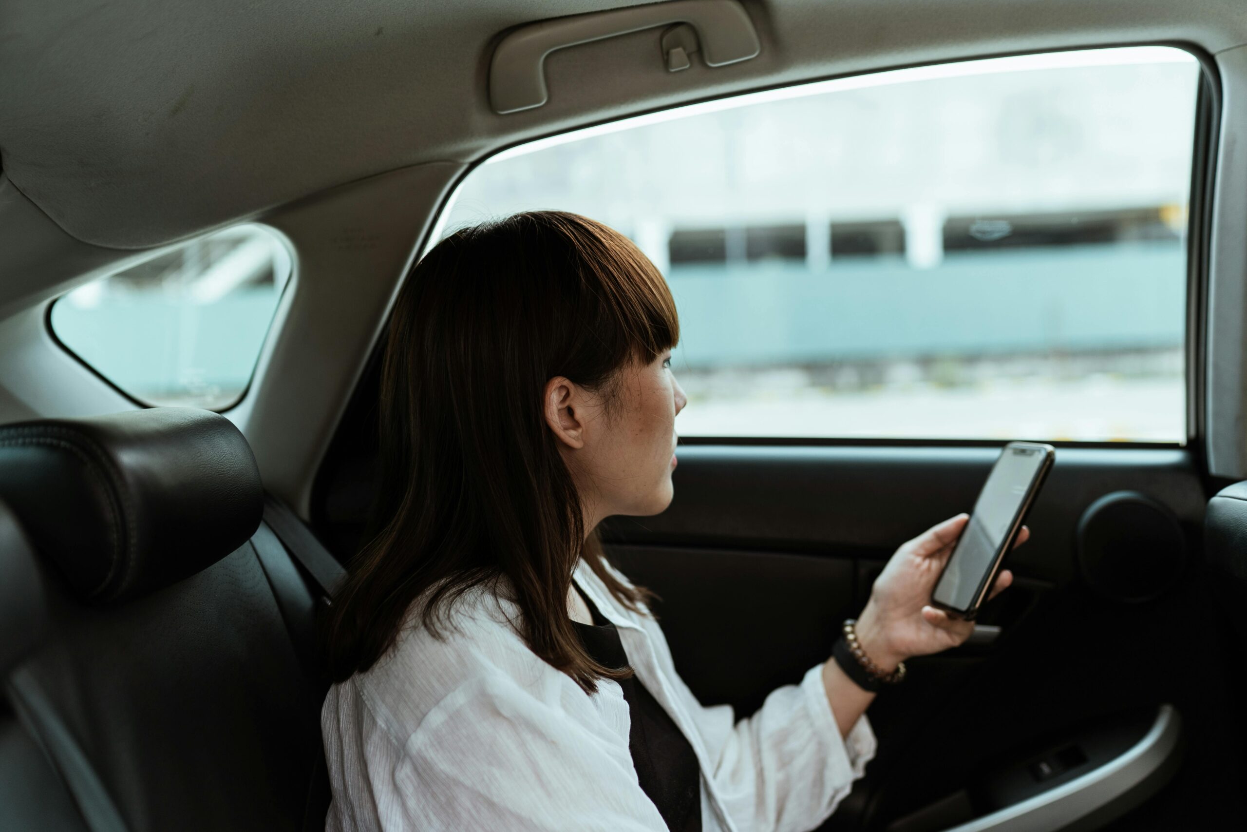 Young woman casually using a smartphone while seated in the backseat of a car, looking outside the window.