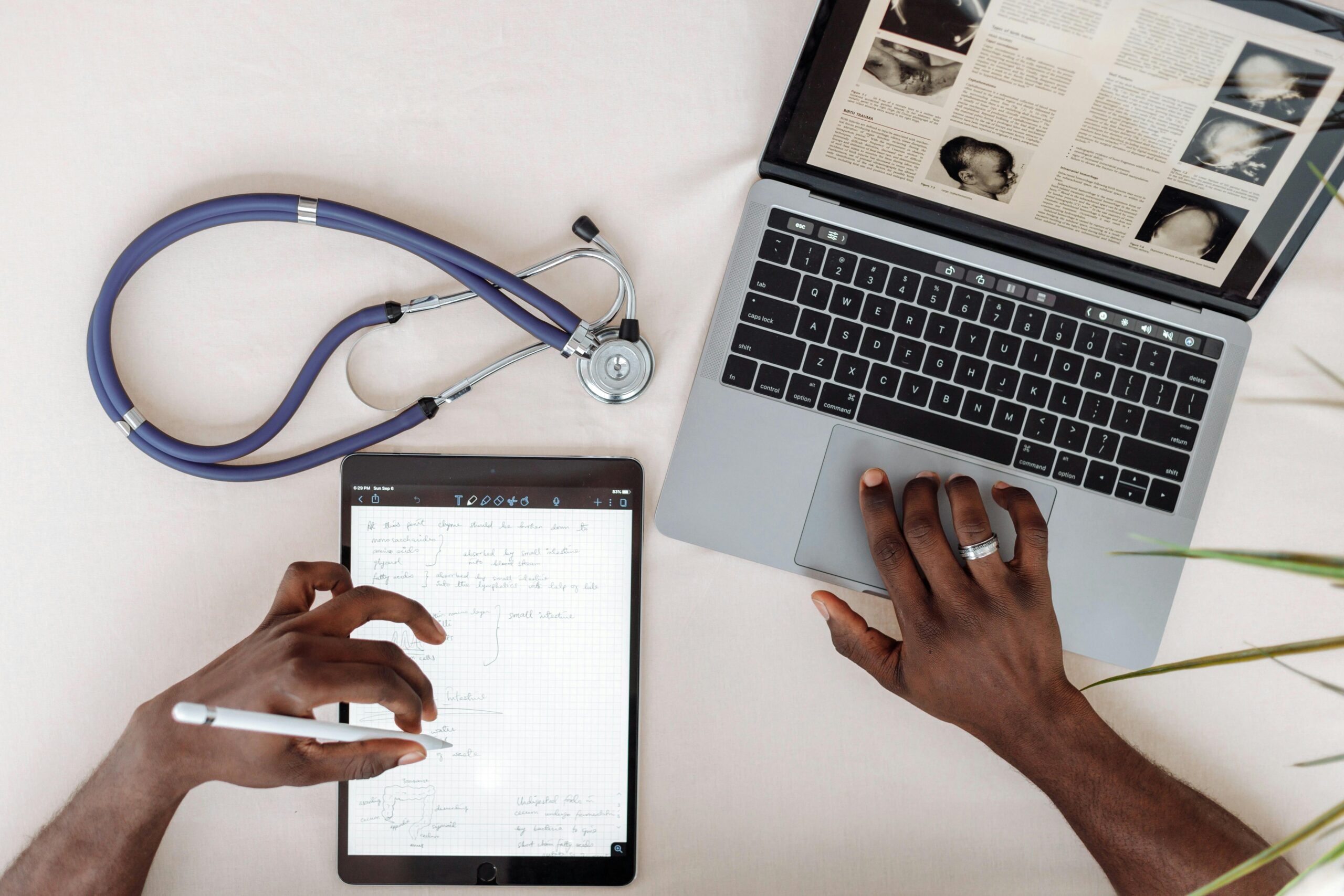 Healthcare professional using tablet and laptop for research with stethoscope on desk.